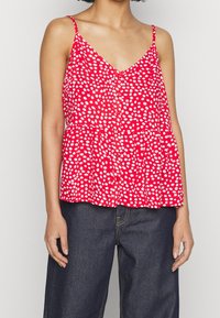 Donna che indossa un top rosso senza maniche con pois bianchi e jeans blu scuro, in piedi contro uno sfondo chiaro e uniforme.