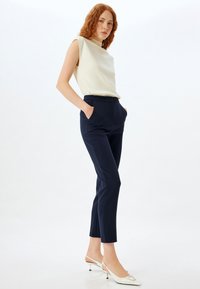 Marineblå skræddersyede bukser med sidelommer og en slim fit, parret med en cremefarvet ærmeløs top. Modellen har hvide slingback hæle på.