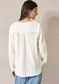 Hvid struktureret bluse med lange ærmer, en detalje med rygplissé og en rund kant, båret med løse blå jeans.