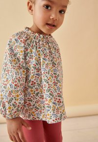 Bloem blouse met geplooide halslijn in het wit, versierd met kleurrijke bloemen en groene bladeren; gecombineerd met roze geribde leggings.