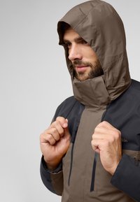 Bruine en zwarte capuchonjas met een hoge kraag, voorzakken met rits en verstelbare trekkoorden zichtbaar. Gladde, weerbestendige stof.