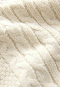 Tissu tricoté couleur crème avec des motifs épais en câble et côtelés, montrant des fibres de laine douces et texturées en gros plan.