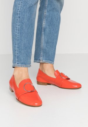 Fødder iført lyse orange læderloafers med sølvfarvede, runde spænde-detaljer kombineret med blå denimjeans mod en ensfarvet baggrund.