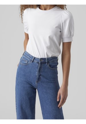 Kobieta w białym t-shircie z bufkami, włożonym do wysokich dżinsów z niebieskiego denimu, na jednolitym szarym tle.