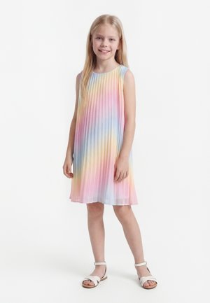 Νέα κοπέλα με μακριά ξανθά μαλλιά, που φοράει αμάνικο, πλισέ φόρεμα σε παστέλ ουράνιου τόξου χρώματα και άσπρα σανδάλια, στέκεται πάνω σε λευκό φόντο.