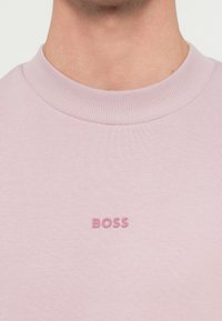 Detailný záber na osobu v svetloružovom pulóvri s okrúhlym výstrihom, na ktorom je na hrudi vyšitý nápis "BOSS" v rovnakom odtieni ružovej farby.