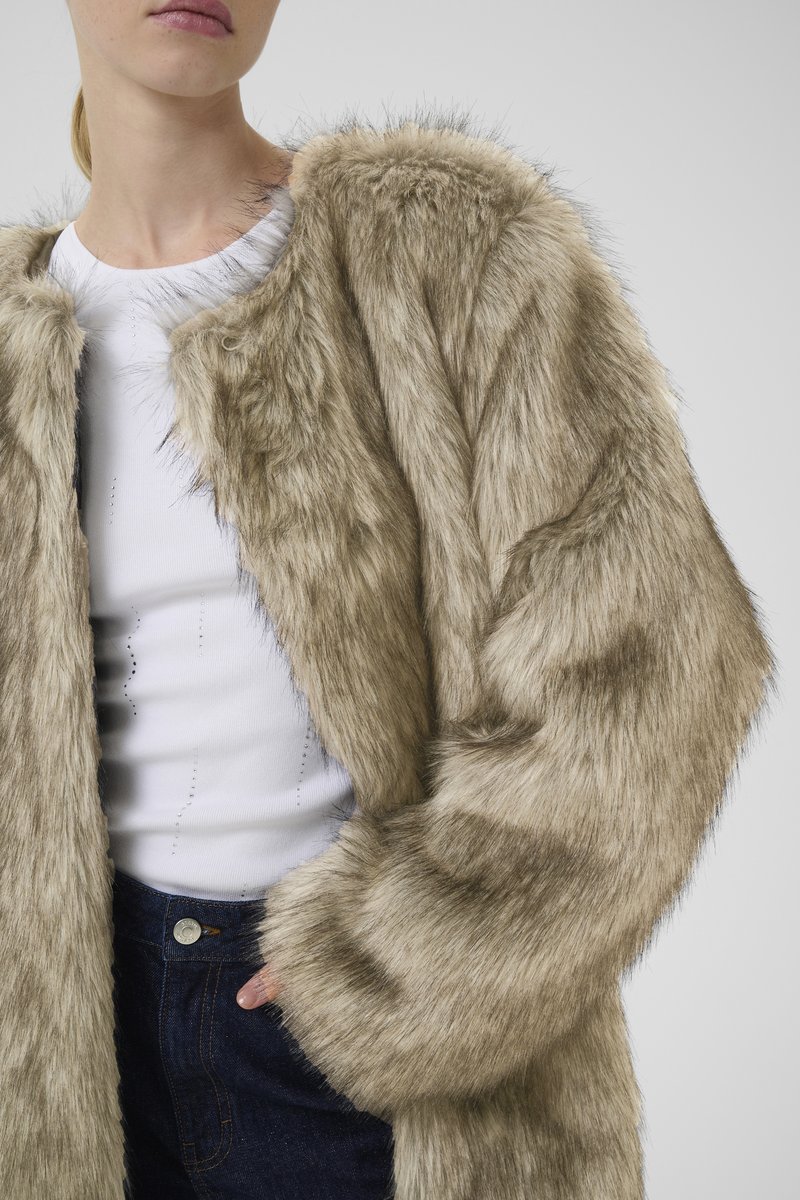 Faux-Fur-Mantel in hellem Beige mit einer sanften Textur, rundem Halsausschnitt und lockerer Passform. Getragen über einem weißen Oberteil und blauen Jeans.