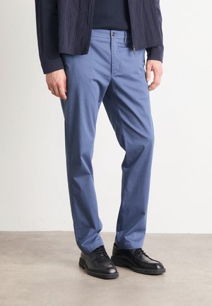 Mand iført blå tætsiddende bukser, sorte snørestøvler og en mørk lynlåsjakke, der står på et lyst gulv.