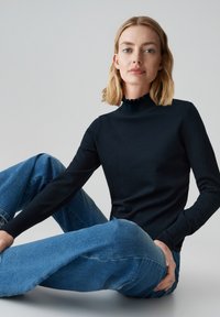 Zwarte langekous top met een hoge gerimpelde halslijn. Gemaakt in combinatie met lichtblauwe high-waisted jeans. Glad textuur, aansluitend ontwerp.