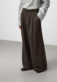 Pantaloni a gamba larga in corduroy marroni con pieghe frontali, vita alta e tasche laterali, abbinati a una camicia a righe con bottoni e scarpe nere.