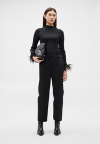 Sieviete, valkājot melnu ribotu topu ar spalvām piedurknēs, melnas pielāgotas bikses un melnas smailas zābaki, rokās turot melnu clutch somiņu.