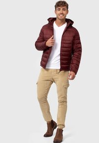 Geacă căptușită burgundy cu glugă, închidere cu fermoar și buzunare laterale, asociată cu pantaloni cargo bej și botine maro cu gleznă. Maiou alb dedesubt.
