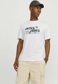 Camiseta blanca de manga corta con un estampado gráfico en color que presenta "JACK & JONES" en azul y negro, combinada con pantalones cortos tipo cargo beige.