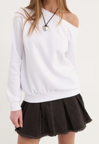 Sudadera blanca off-shoulder con dobladillo elástico, combinada con una falda acampanada de mezclilla oscura con un dobladillo deshilachado. Colgante en forma de corazón sobre un cordón negro.