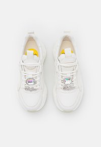 White sneakers with a textured leather upper, rounded shape, thick sole, and a shiny decorative emblem. Yellow insoles visible inside.