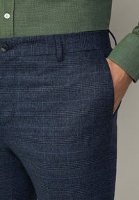 Donkerblauwe geruite broeken van een gestructureerde stof, met een gladde voorkant, zijzakken en een knoopsluiting. Groene shirt zichtbaar erboven.