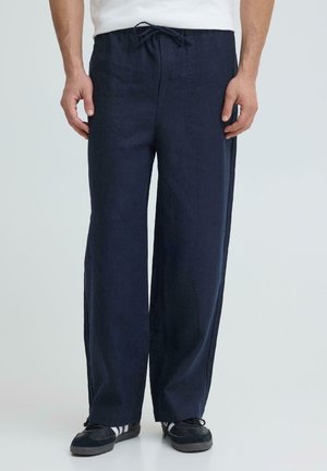 Mann trägt locker sitzende marineblaue Hose mit Kordelzugbund und schwarze Turnschuhe mit weißen Streifen.