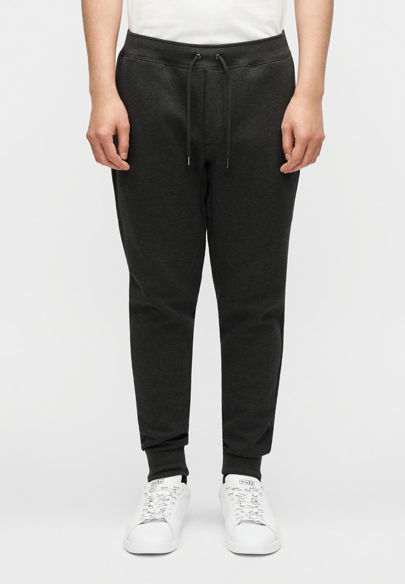 Σκούρο γκρι παντελόνι jogger με ελαστική μέση και κορδόνι. Διαθέτει ριμπ μανσέτες και λεία υφή από βαμβακερό μείγμα. Φοριέται με λευκά αθλητικά παπούτσια.