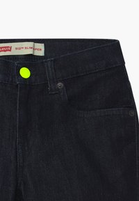 Dark denim jeans featuring a slim fit with a bright yellow button accent, contrasting rivets, and classic five-pocket design.