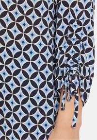 Tissu bleu et marine à motifs géométriques ; présente une manche froncée avec des liens pour un ajustement réglable ; texture lisse.