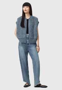 Canotta senza maniche di colore blu chiaro, realizzata in maglia con dettagli in peluche testurizzati, indossata sopra una maglietta nera e abbinata a jeans a gamba larga blu chiaro. Scarpe nere slip-on.