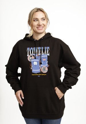 Uśmiechnięta kobieta w czarnej bluzie z kapturem z kreskówkowymi ręcznikami i napisem „Towelie” oraz „Nie mam pojęcia, co się dzieje” na białym tle.