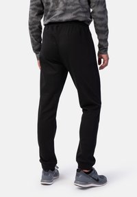 Schwarze Jogginghosen mit einem elastischen Bund, schmal zulaufenden Beinen und einer weichen Textur. Tragen mit grauen Sportschuhen, von hinten betrachtet.