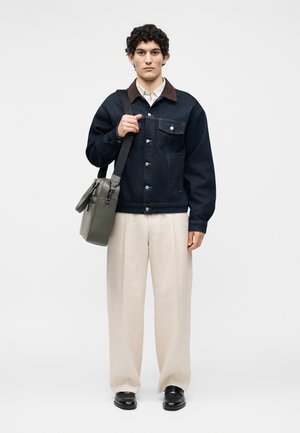 Fiatal férfi sötét farmerdzsekit és világos bézs nadrágot visel, szürke válltáskát tart, homogén fehér háttér előtt áll.