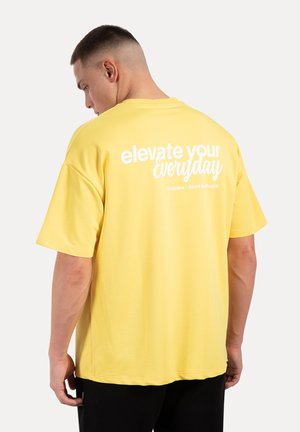 Gelbes Baumwoll-T-Shirt mit lockerer Passform, mit einem weißen Druckslogan "elevate your everyday" auf dem Rücken. Kurze Ärmel und Rundhalsausschnitt.