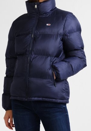 Doudoune bleu marine avec col montant et fermeture éclair à l'avant, portée par une personne avec une main dans la poche et un jean foncé visible.