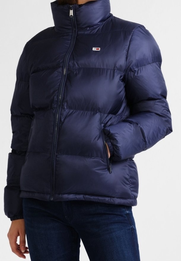 Doudoune bleu marine avec col montant et fermeture éclair à l'avant, portée par une personne avec une main dans la poche et un jean foncé visible.