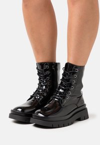 Bottes courtes en cuir verni noir avec une plateforme noire épaisse, des œillets argentés et un design à lacets. Texture lisse avec une finition brillante.