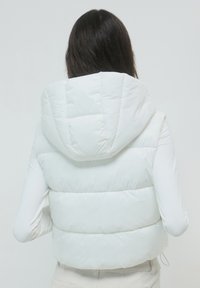 Donna vista di spalle che indossa un gilet imbottito bianco con cappuccio sopra una maglia a maniche lunghe, su uno sfondo chiaro e semplice.