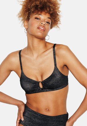 Femme aux cheveux bouclés portant un haut de bikini noir pailleté avec un détail en forme de trou de serrure et un bas taille haute assorti, posant avec la main sur la hanche.