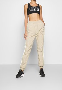 Svart croptop med "LEVI'S"-logotyp, beige joggingbyxor med vita sidostripor och elastiska muddar, tillsammans med svarta sportskor.