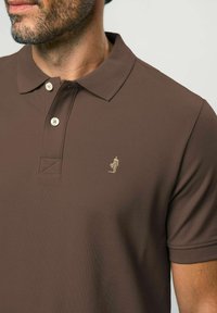 Tricou polo maro din țesătură texturată, cu guler clasic, trei nasturi și un mic logo brodat pe piept.