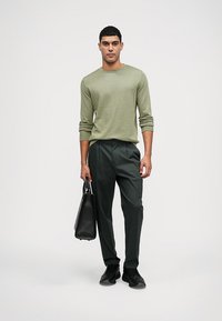 Zelená pletená košeľa s dlhými rukávmi v kombinácii s tmavými nohavicami. Čierna kožená taška držaná v pravej ruke. Čierne tenisky dotvárajú outfit.