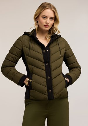 Frau trägt olivgrüne gesteppte Jacke mit schwarzem Rand, schwarzen Knöpfen und goldenen Schmuckstücken, steht mit in den Taschen gesteckten Händen.