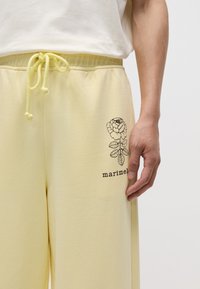 Dzeltenie kokvilnas sporta bikses ir ar elastīgu jostu un auklu, un uz kreisā kājas ir ziedu grafika ar tekstu "marimekko".