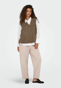 Bruin gebreid vest met V-hals en knopen, gedragen over een wit shirt, gecombineerd met lichtbeige broek en zwarte instappers.