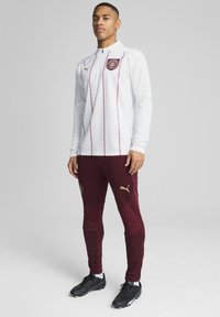 Balta ilgomis rankovėmis sportinė maikutė su raudonmedžio akcentais ir auksu logotipu, derinama su raudonmedžio aptemptomis kelnėmis su auksu logotipu ir juodais batais.