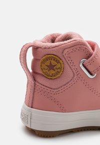 Bota de cuero rosa con cuello de tela texturizada, suela de goma y parche circular en tono marrón claro con un logo de estrella. Incluye ojetes de metal y detalles de costura.