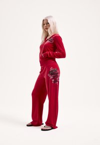 Červený zamatový teplákový komplet so zapínanou bundou na zips a nohavicami so širokými nohavicami. Na bunde a bokoch sú čierne grafické nápisy, komplet je doplnený žabkami.