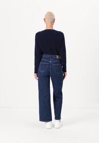 Marineblauwe geribbelde trui en hoge blauwe jeans. De jeans hebben een klassieke snit met achterzakken en een tan leren label. Witte sneakers.