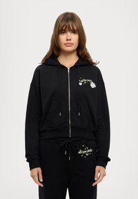 Črna zip-up kapuca z žepom spredaj, ki vključuje majhen barvit grafični dizajn na levi strani prsi. Mehka tkanina, udoben kroj.