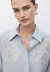 Primer plano de una mujer que lleva una blusa transparente de color azul claro con patrones florales bordados en el pecho y pendientes de aro plateados.