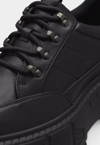 Μαύρο δερμάτινο αθλητικό παπούτσι με παχιά σόλα, ελαστικά μαύρα κορδόνια στερεωμένα με μεταλλικά οπές και ραμμένες λεπτομέρειες πάνελ στο πλάι.