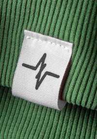 Grüner gerippter Stoff mit einem weißen Etikett, das ein schwarzes Herzschlag-Graphic zeigt. Texturierte Nahaufnahme des Materials und der Etikettendetails.