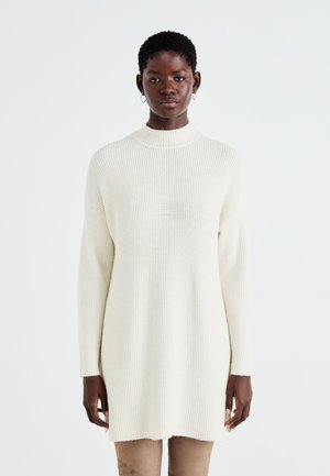 Robe pull en tricot crème épaisse à col montant, manches longues et texture côtelée, offrant une coupe décontractée et des fentes sur les côtés.