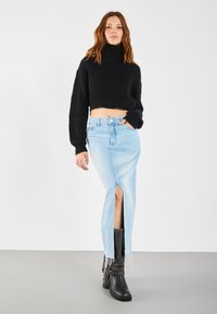 Maglione nero corto con texture a coste, abbinato a una gonna di jeans leggera blu chiaro a vita alta con uno spacco frontale. Stivali neri con tacco alla caviglia.